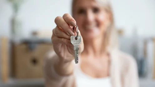 Junge Frau hält Haustürschlüssel in die Höhe und übergibt damit das verkaufte Haud an den Käufer.