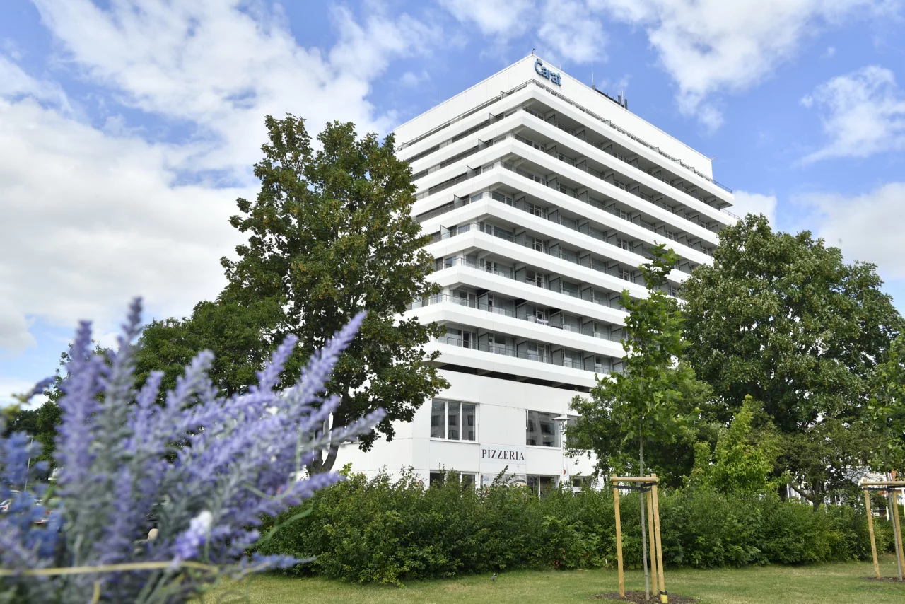 Ein weißes großes Hotel auf einer grünen Wiese und grünen Baumen rundherum und blauem Himmel