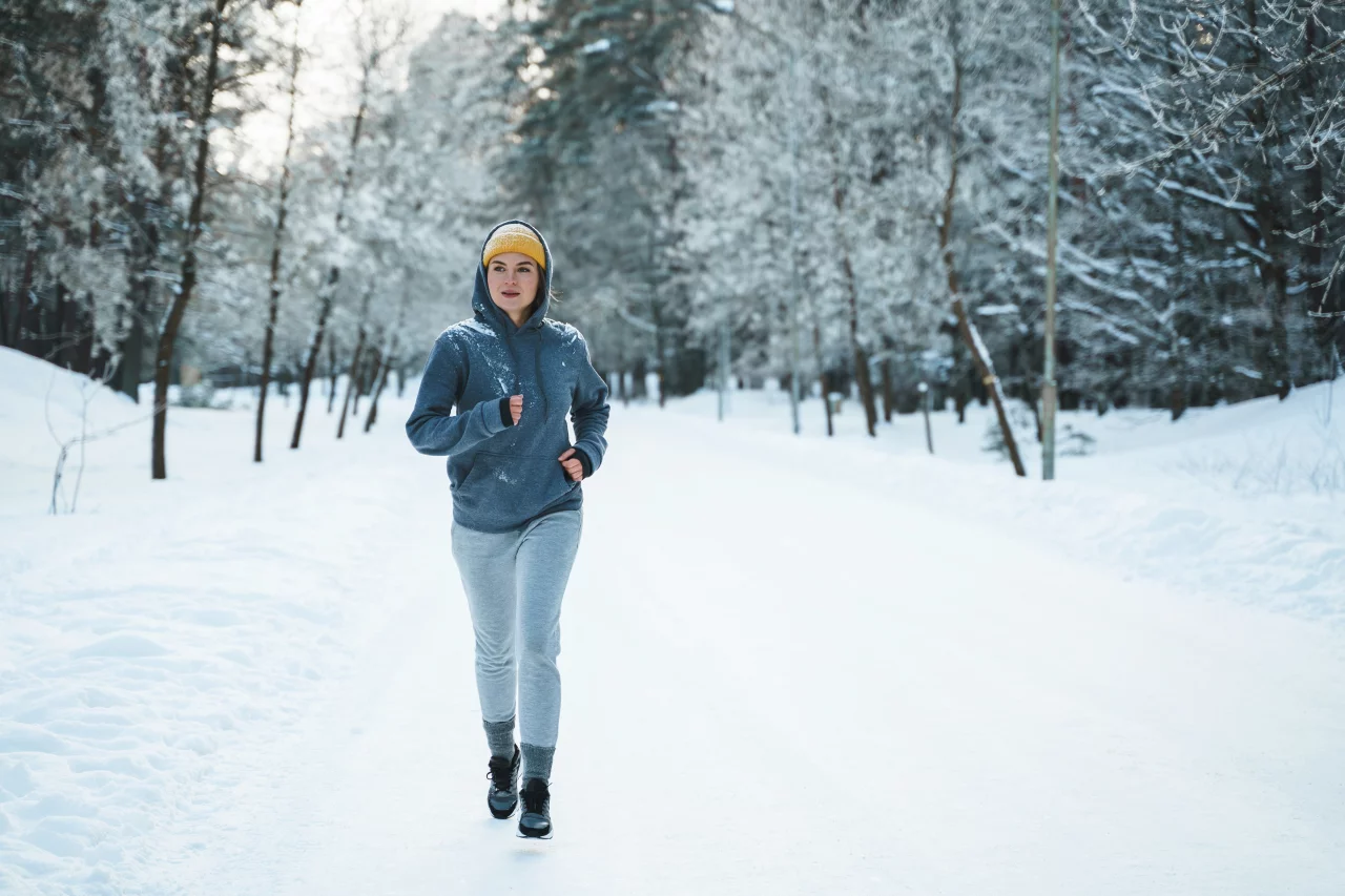 Eine Frau mit grauen Jogging-Sachen und einer gelben Wollmütze läuft durch einen schneebedeckten Wald