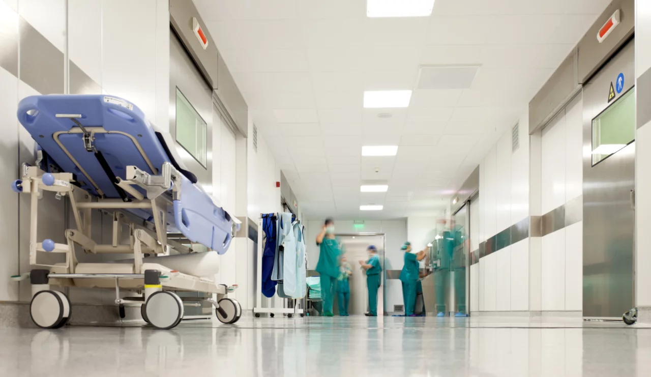 Ein Krankenhaus Flur. Am Ende sieht man Pflegerinnen und im Vordergrund ein leeres blaues Bett.
