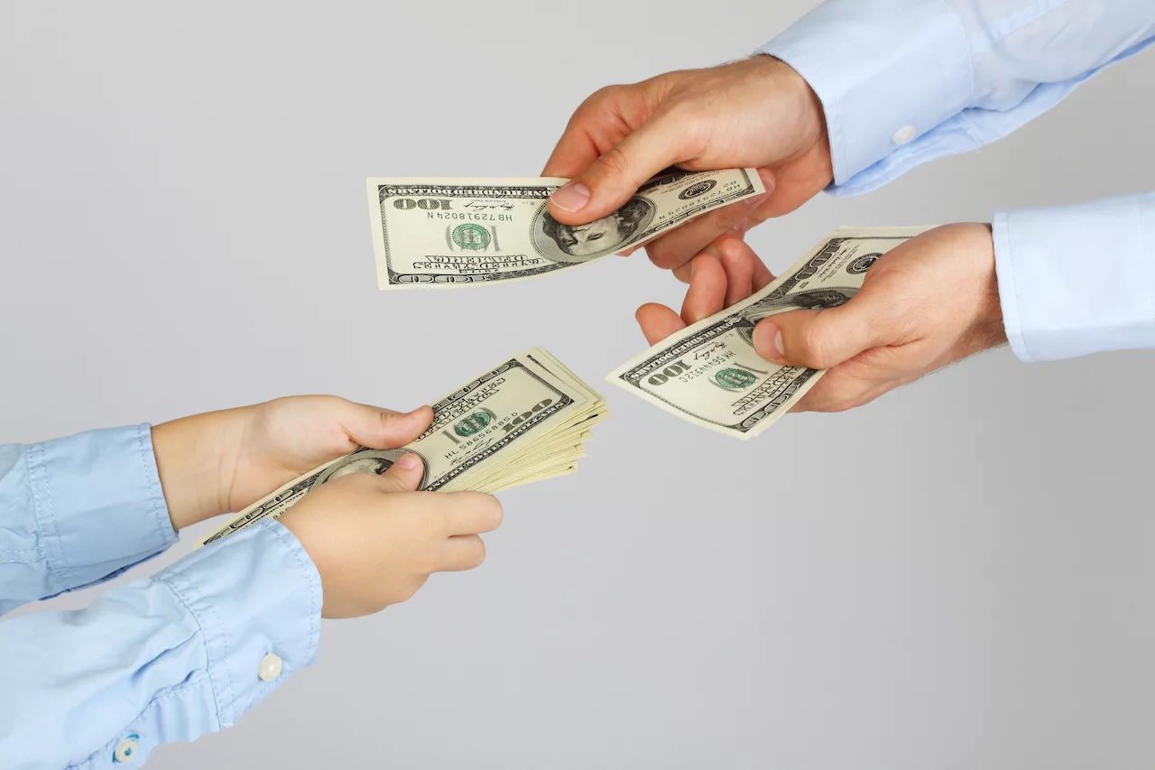 Männerhände geben den Jungenhänden Geld in Form von Hundert-Dollar-Scheinen. Geschäftsleute geben Geschäftsleuten Geld. Vater und Sohn.