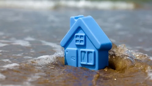 Überschwemmung, Starkregen, Bild von blauem Spielzeug-Plastikhaus auf dem Sand, das von Wellen umspült wird, Symbolbild