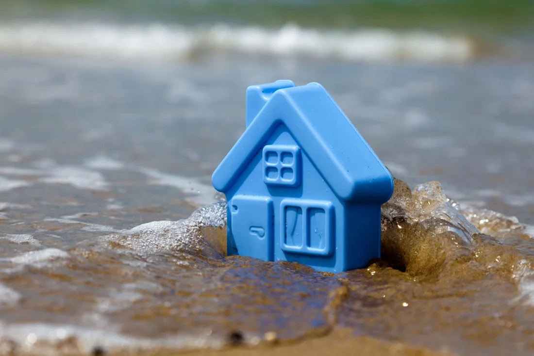 Überschwemmung, Starkregen, Bild von blauem Spielzeug-Plastikhaus auf dem Sand, das von Wellen umspült wird, Symbolbild