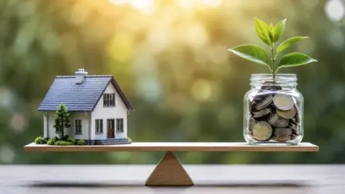 Modellhaus und mit Münzen gefülltes Sparglas mit Pflanze liegen auf einer Waage im Gleichgewicht.