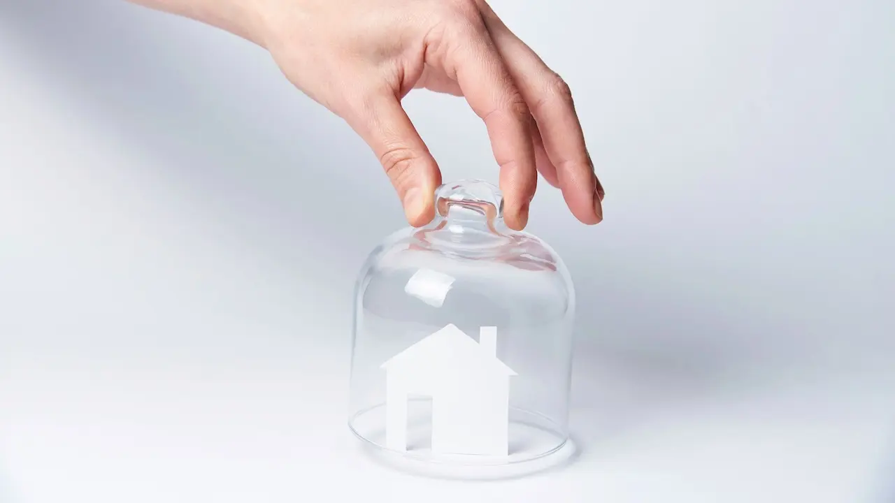 Hand steckt ein Papierhaus unter einen Glasdeckel - Symbol für Mietendeckel.