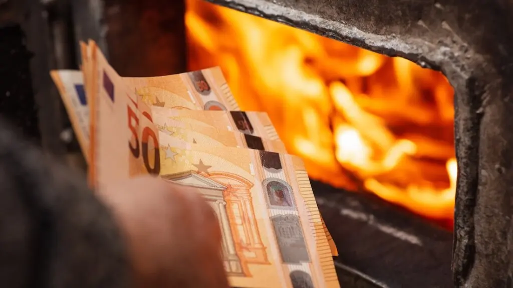Euro-Scheine in der Hand eines Mannes neben dem Heizkessel, dessen Türen offen stehen und aus dem Flammen lodern. Hohe Ausgaben für die Beheizung des Hauses, teure Heizkosten