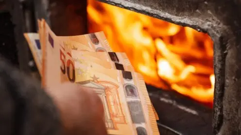 Euro-Scheine in der Hand eines Mannes neben dem Heizkessel, dessen Türen offen stehen und aus dem Flammen lodern. Hohe Ausgaben für die Beheizung des Hauses, teure Heizkosten