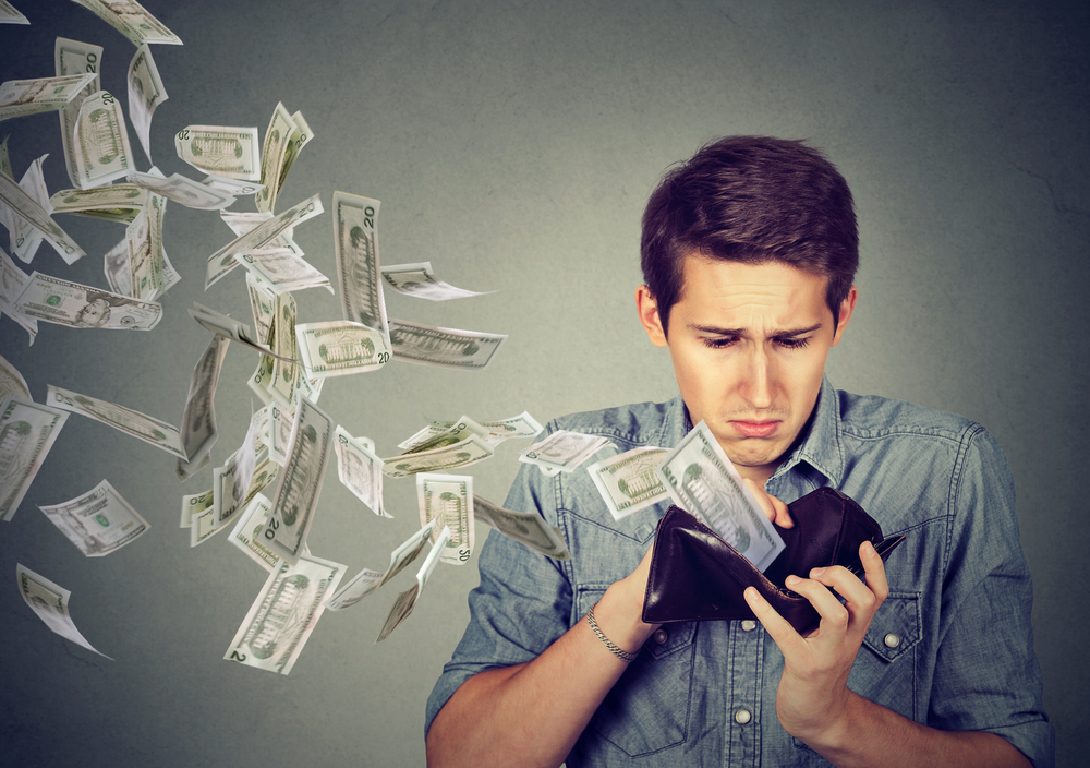 Money Depletion Mann guckt traurig auf die Brieftasche in seiner Hand, aus der zahlreiche Geldscheine herausfliegen.