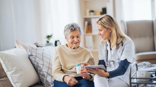 Eine ältere Frau sitzt auf einer Couch und eine Krankenschwester reicht ihr ein Getränk