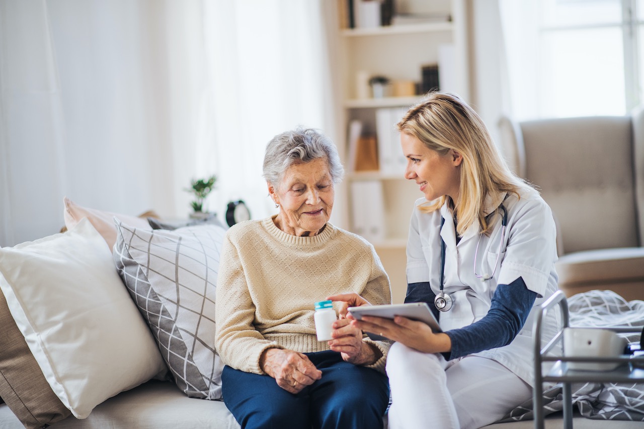 Eine ältere Frau sitzt auf einer Couch und eine Krankenschwester reicht ihr ein Getränk