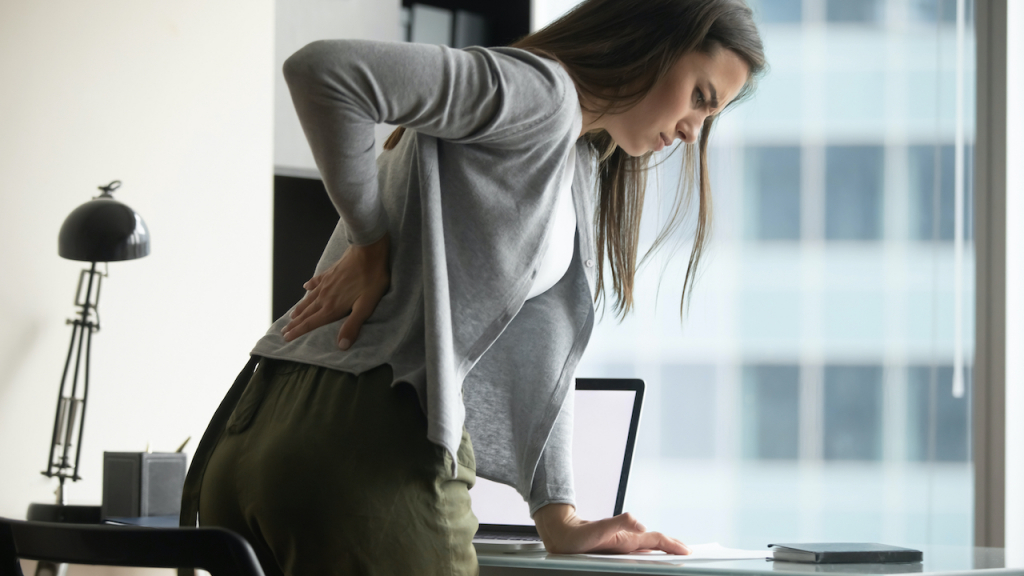 Junge FRau, dei vom Schreibtisch mit Rückenschmerzen aufsteht