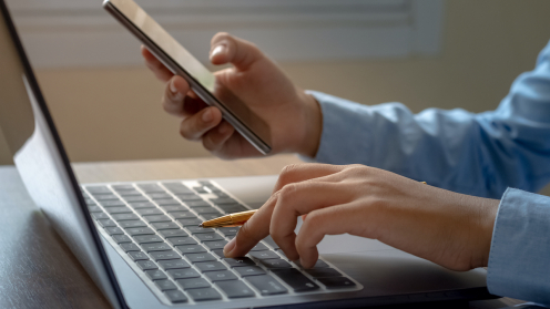 Ein Laptop an dem jemand sitzt, man sieht nur die Hände. Eine tippt und einer anderen hält der Mann ein Handy