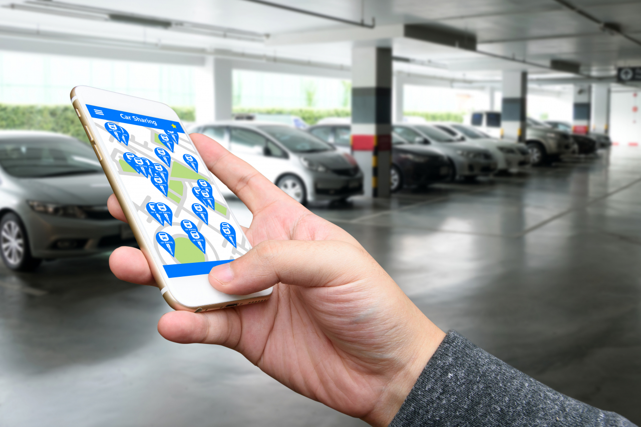 Ein Handy wird in der Hand gehalten mit einer Car Sharing App geöffnet. Im Hintergrund sieht man Auto in einem Parkhaus