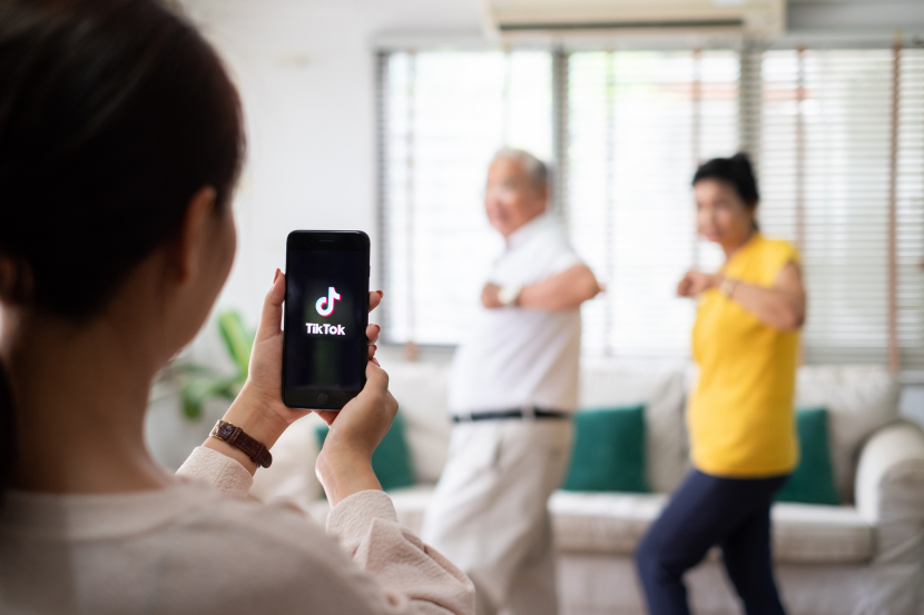 Eine junge Frau im Vordergrund von hinten gefilmt hält ein handy hoch, darauf ist die App TikTok geöffnet. Sie filmt ein älteres Ehepaar die eine Choreografie machen