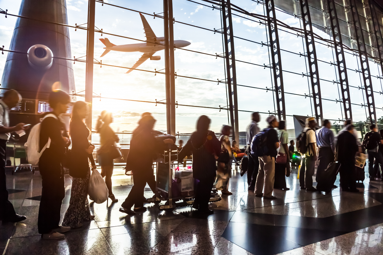 Eine Warteschlange in einer Flughafen-Halle vor einem großen Fenster durch das man ein Flugzeug abheben sieht