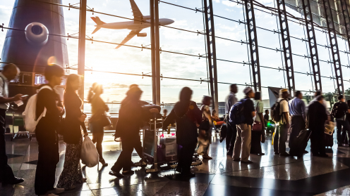 Eine Warteschlange in einer Flughafen-Halle vor einem großen Fenster durch das man ein Flugzeug abheben sieht