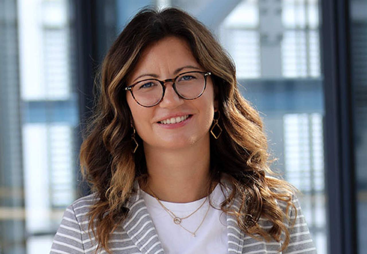 Janine Spieker, junge Frau mit Brille und schulterlangem, braunen, leicht gelocktem Haar, lächelt in die Kamera.