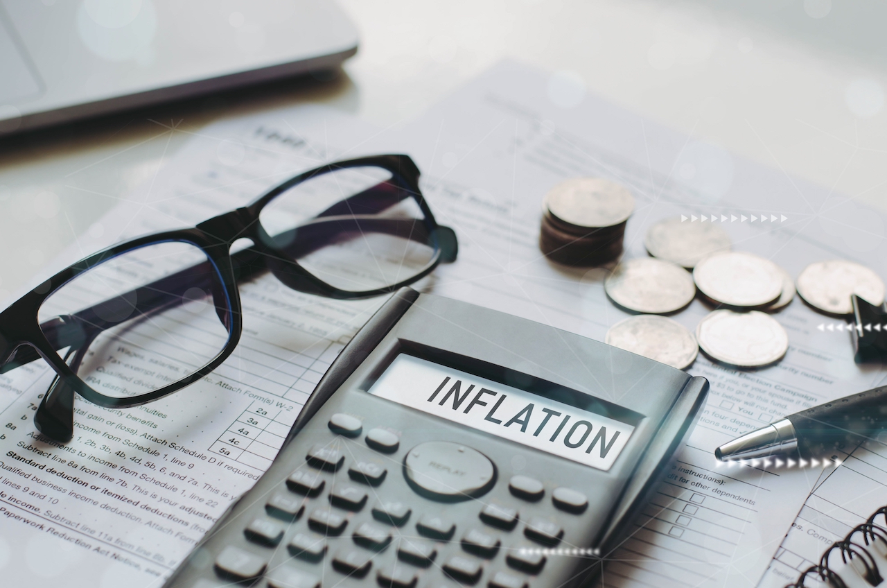 Ein Taschenrechner liegt auf einem Blatt Papier und zeigt den Begriff Inflation. Um ihn liegen eine Brille, Muenzen und eine Kugelschreiber.Auf dem Schreibtisch l