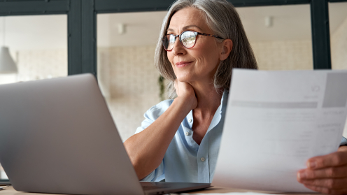 Eine Frau mit grauen Haaren und Brille sitzt an ihrem Laptop und hält ein Schreiben in der Hand