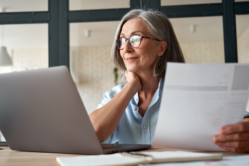 Eine Frau mit grauen Haaren und Brille sitzt an ihrem Laptop und hält ein Schreiben in der Hand