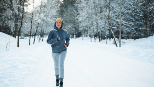 Eine Frau mit grauen Jogging-Sachen und einer gelben Wollmütze läuft durch einen schneebedeckten Wald