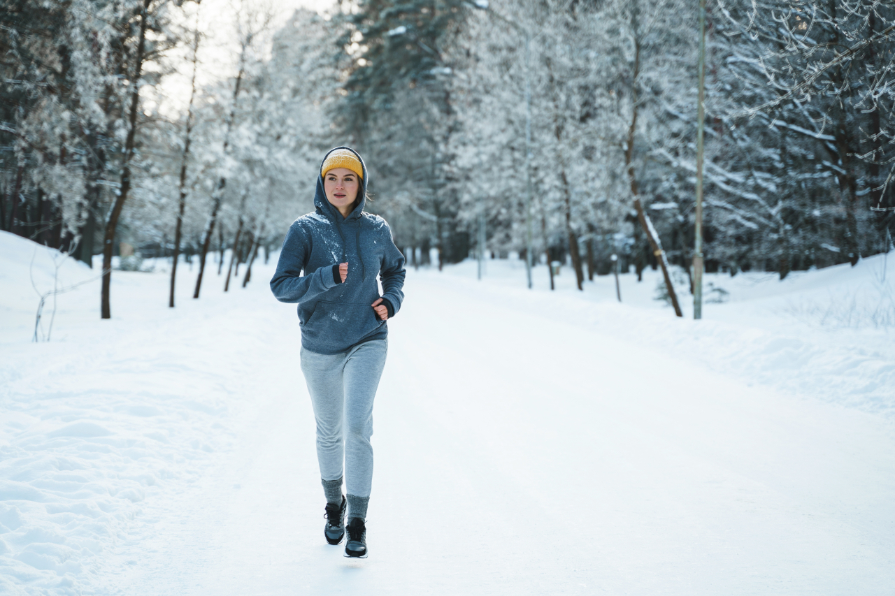 Eine Frau mit grauen Jogging-Sachen und einer gelben Wollmütze läuft durch einen schneebedeckten Wald