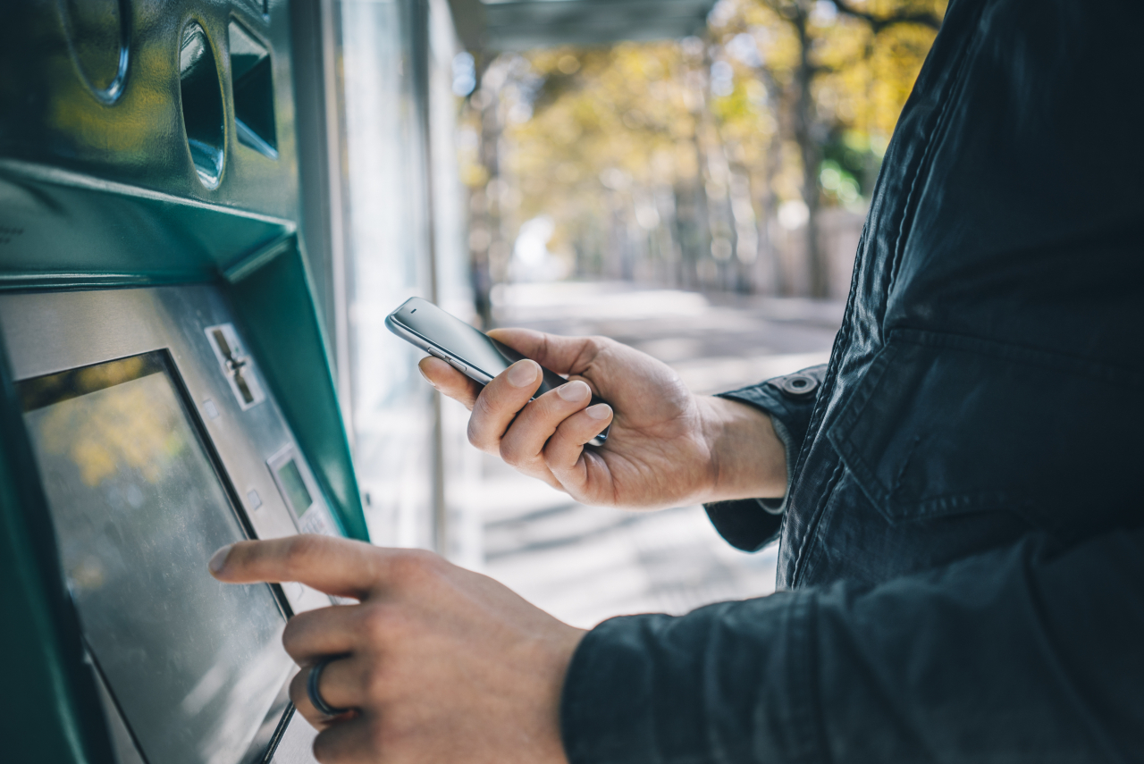 Ein Mann tippt an einem Geldautomaten und hält in der anderen Hand sein Handy