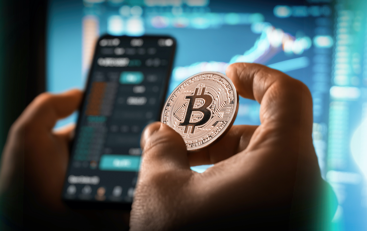 Jemand hält eine Bitcoin-Münze vor einem Smartphone und einem Computerbildschirm.