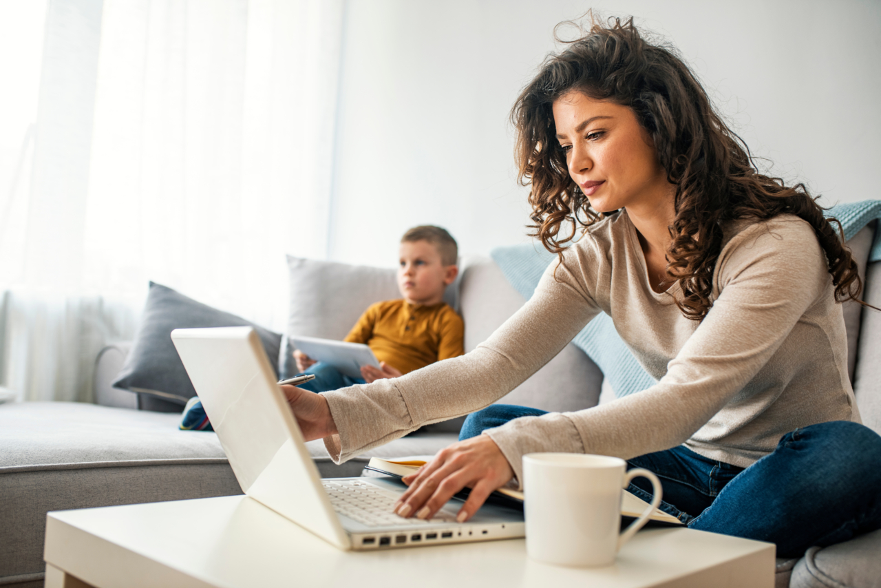 Eine Frau sitzt vor dem Laptop hinter ihr auf der Couch sitzt ein kleiner Junge