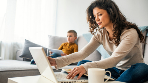 Eine Frau sitzt vor dem Laptop hinter ihr auf der Couch sitzt ein kleiner Junge