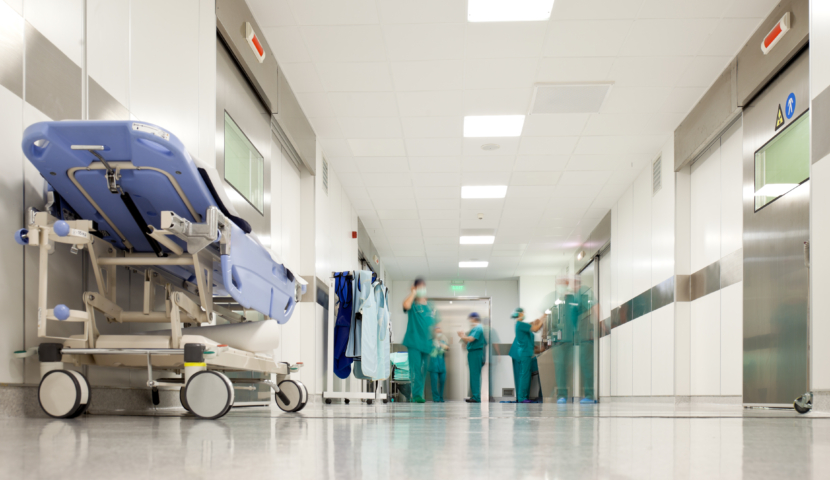Ein Krankenhaus Flur. Am Ende sieht man Pflegerinnen und im Vordergrund ein leeres blaues Bett.