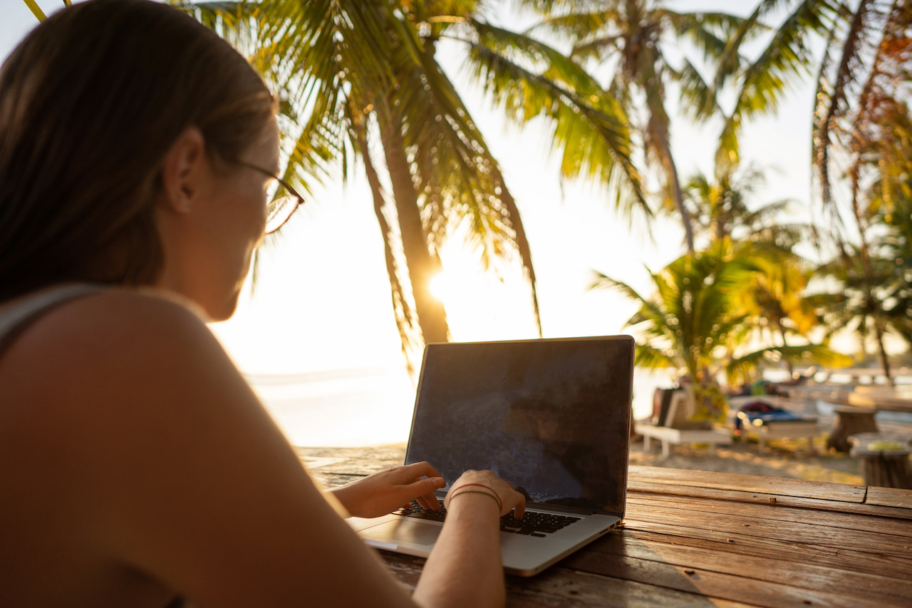 Eine Frau sitzt am Laptop und im Hintergrund Palmen und Sonne