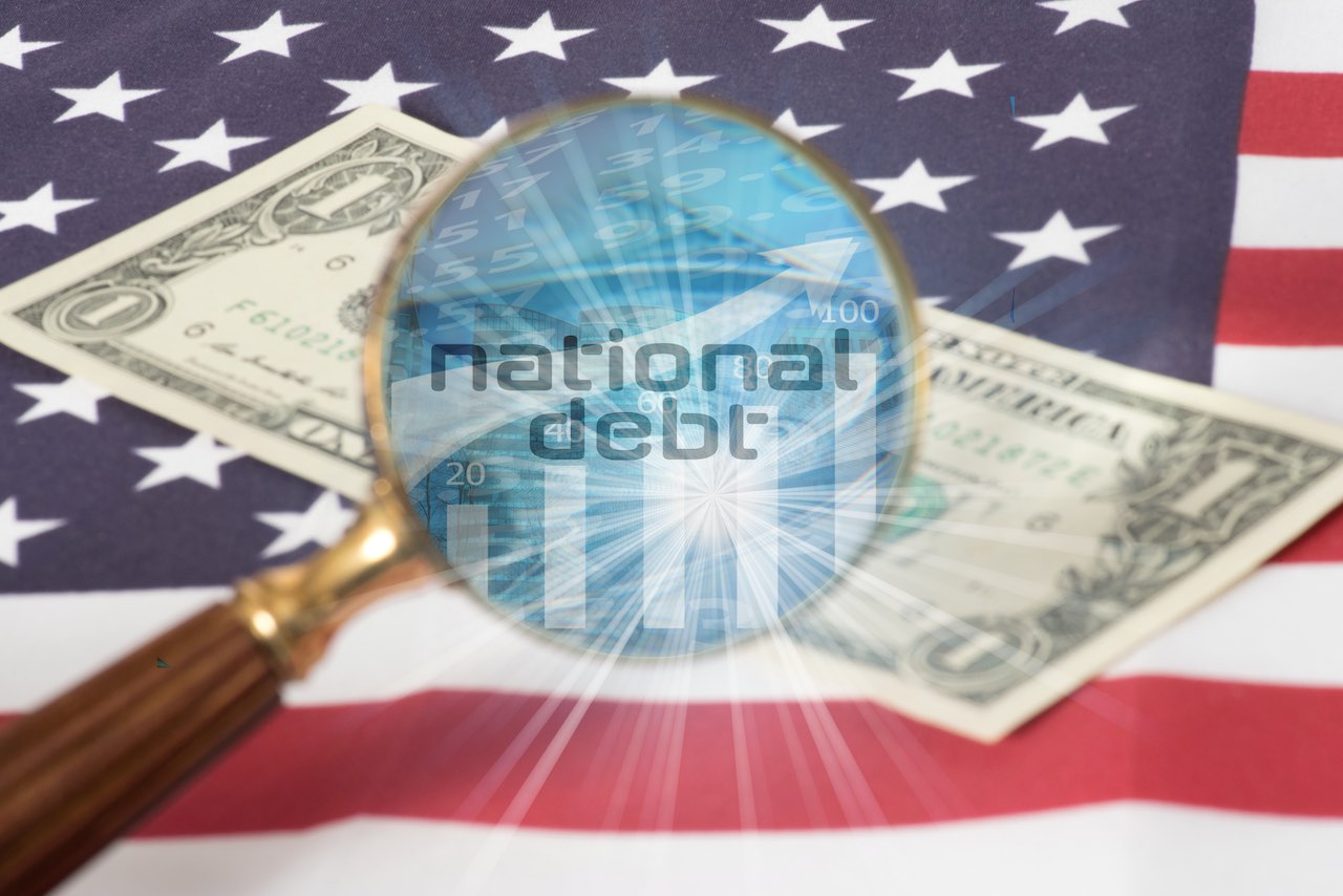 Lupe, durch der Schriftzug "Natonal Debt" platziert auf einer Dollarnote, die auf der US-Flagge liegt, zu sehen ist