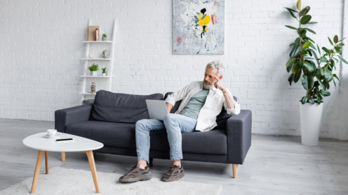 Mann mit grauen Haaren benutzt Laptop, während er auf Couch neben Tasse Kaffee und Smartphone mit leerem Bildschirm auf Couchtisch sitzt