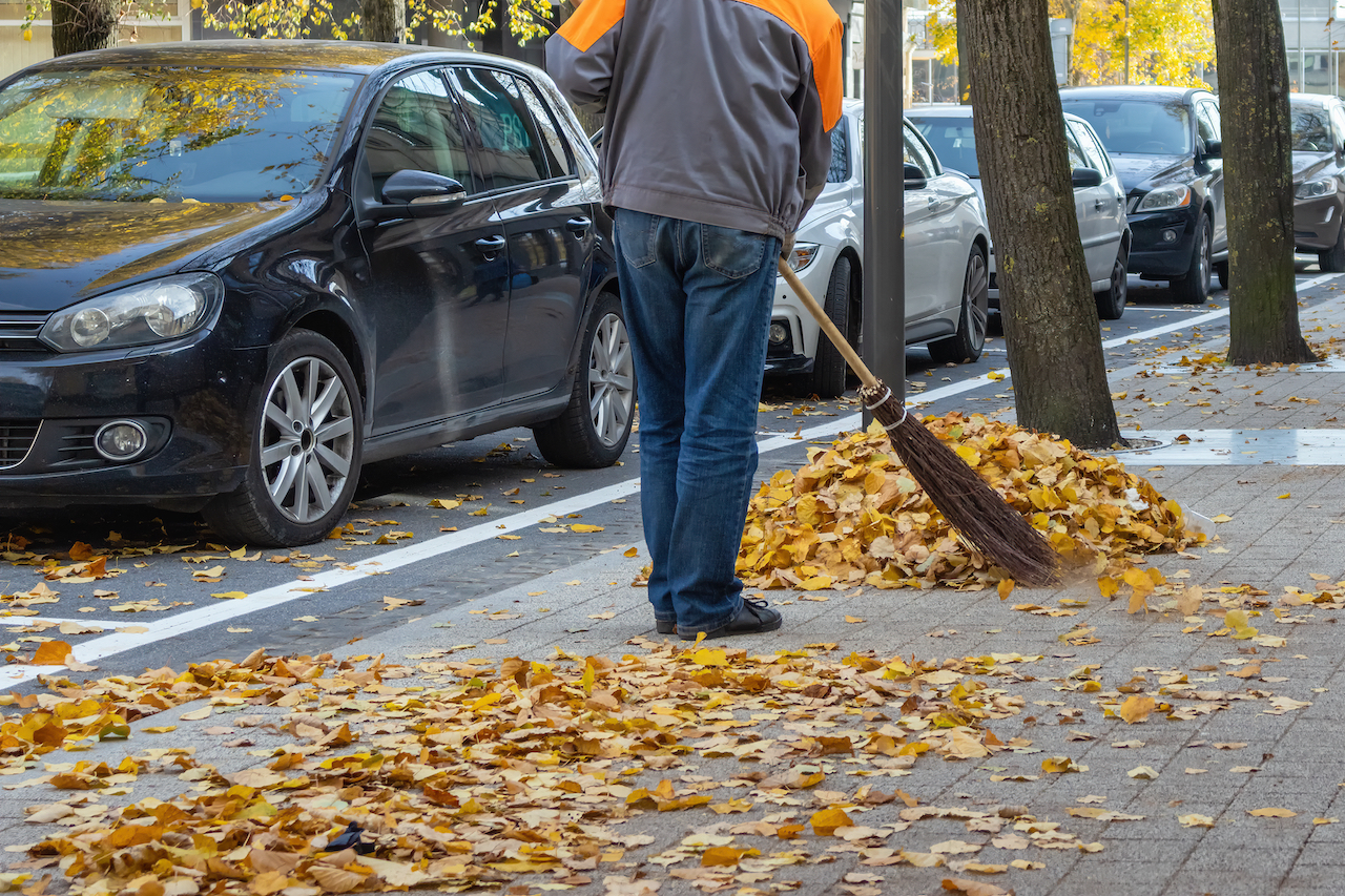 Ein Mitarbeiter der Stadt fegt im Herbst Laub vom Gehweg in einen Haufen.