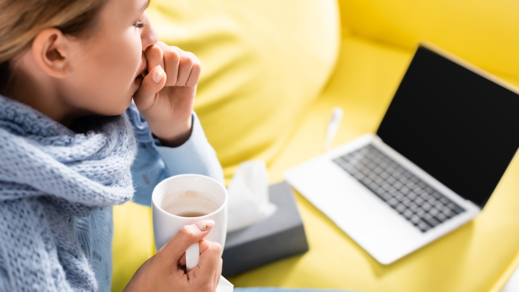 Kranke Frau hustet, während sie Tasse in der Nähe von Laptop und Servietten auf verschwommenem Hintergrund hält