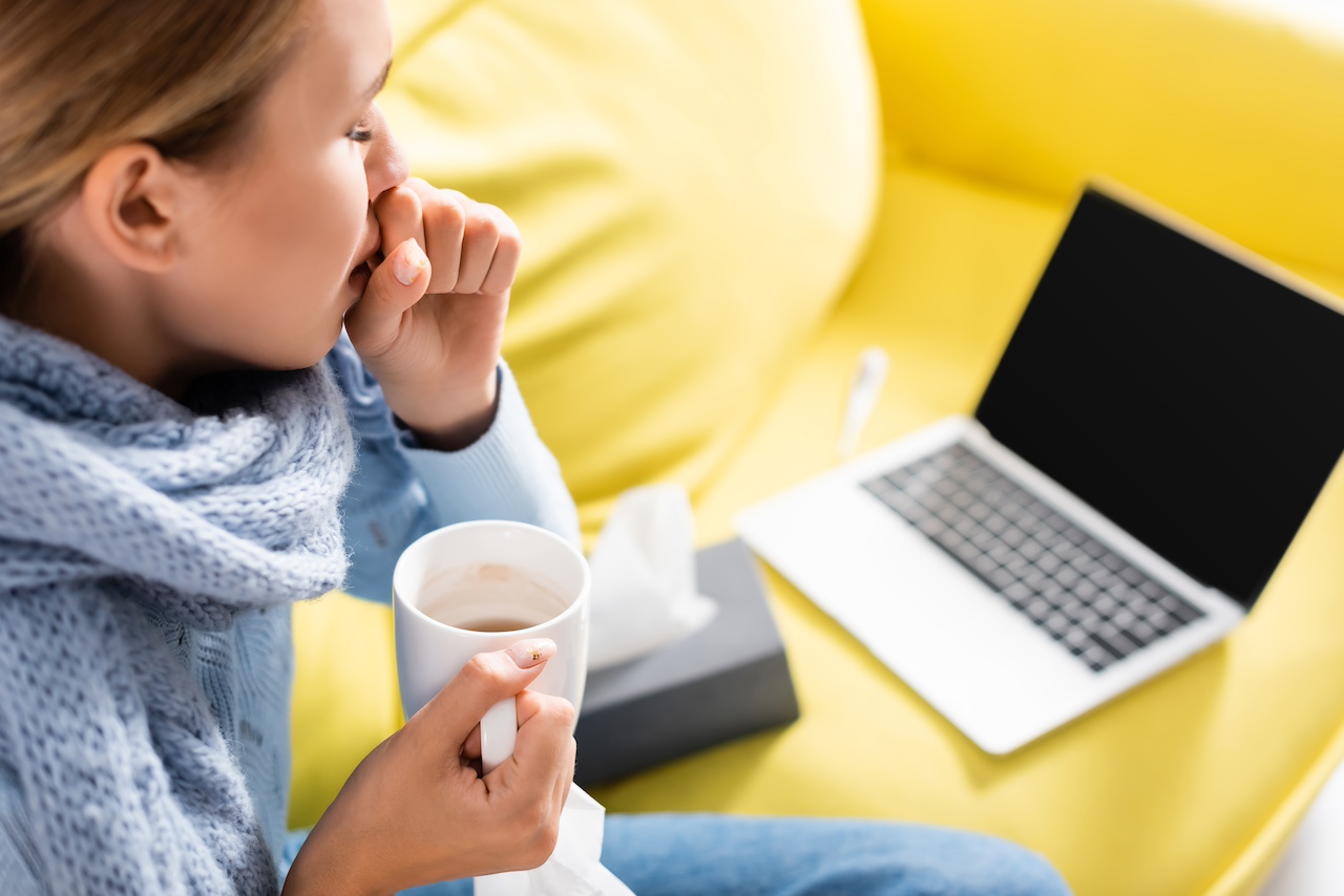 Kranke Frau hustet, während sie Tasse in der Nähe von Laptop und Servietten auf verschwommenem Hintergrund hält