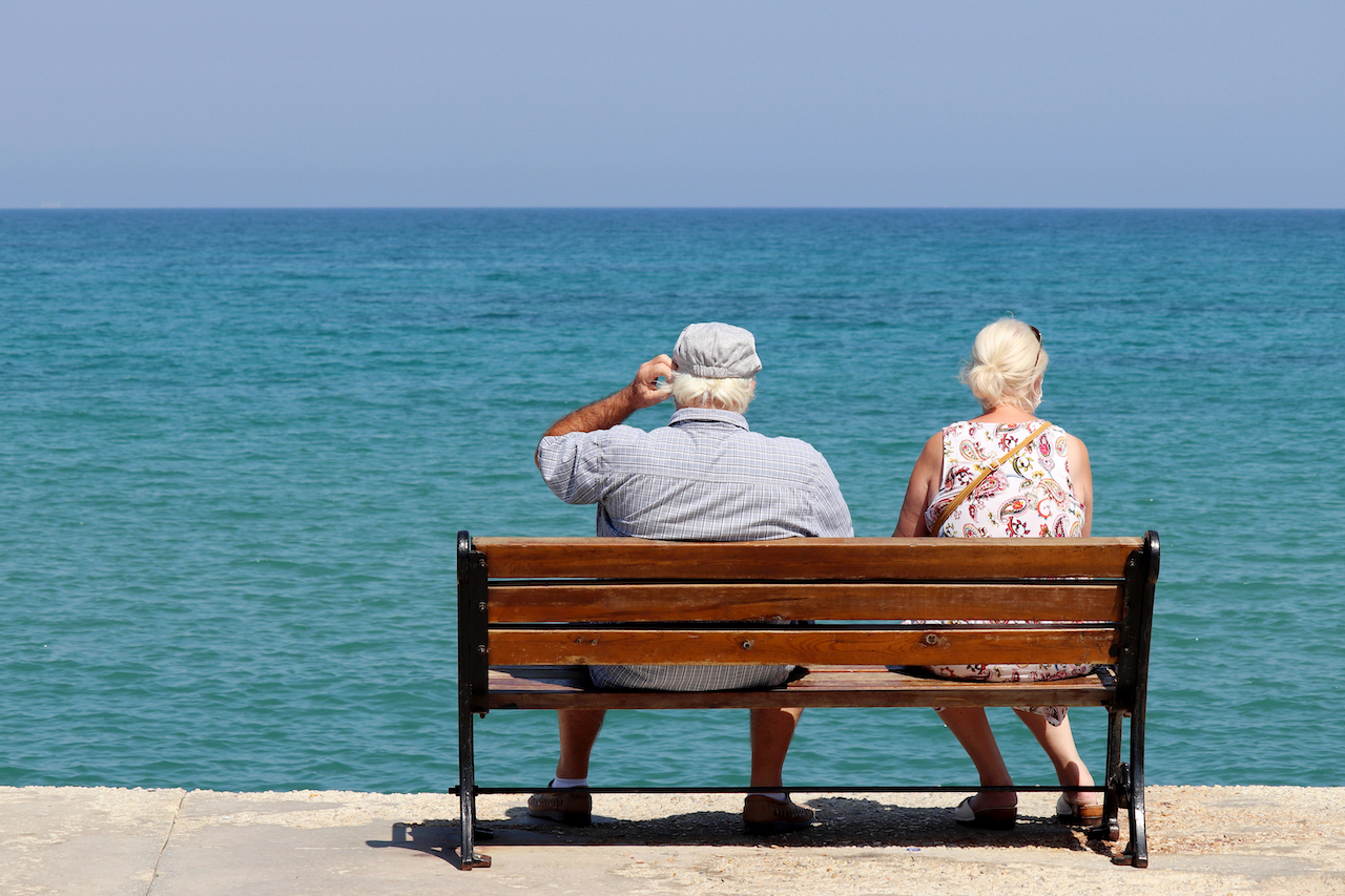Älteres Paar sitzt auf einer Holzbank und blickt aufs Meer, Rückansicht.