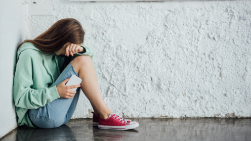 Trauriges weinendes Mädchen sitzt in der Nähe einer Mauer und hält Smartphone in der Hand