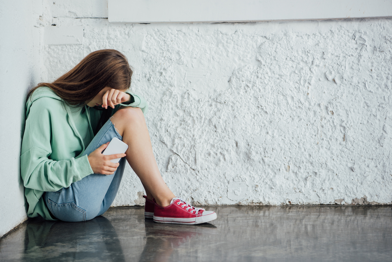 Trauriges weinendes Mädchen sitzt in der Nähe einer Mauer und hält Smartphone in der Hand