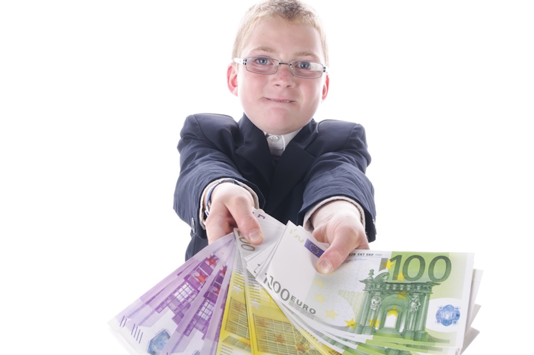 Kleiner Junge im dunklen Anzug spielt mit einem Bündel Geld, isoliert vor weißem Hintergrund.