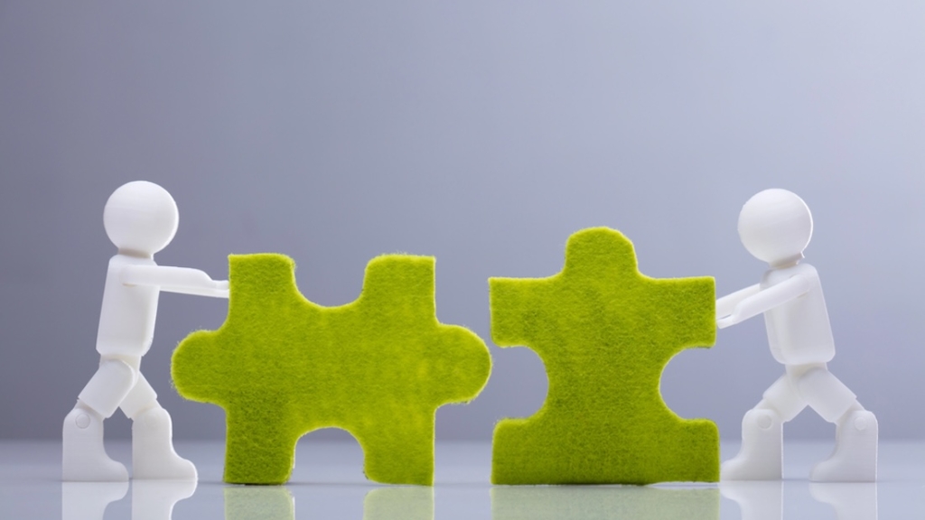 Ansicht von zwei Miniatur-Menschenfiguren, die auf grauem Hintergrund grüne Puzzles lösen