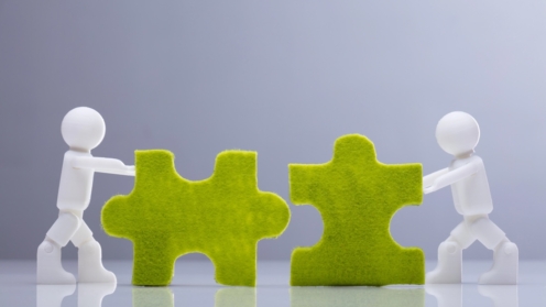 Ansicht von zwei Miniatur-Menschenfiguren, die auf grauem Hintergrund grüne Puzzles lösen
