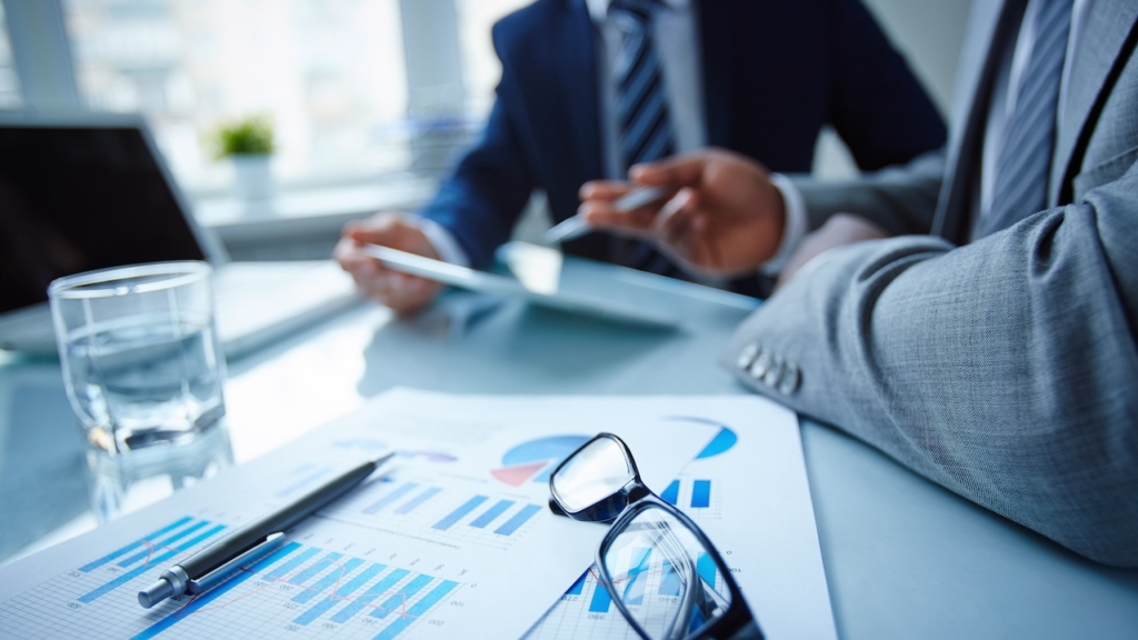 Image of eyeglasses and financial documents at workplace with businessmen discussing ideas near by