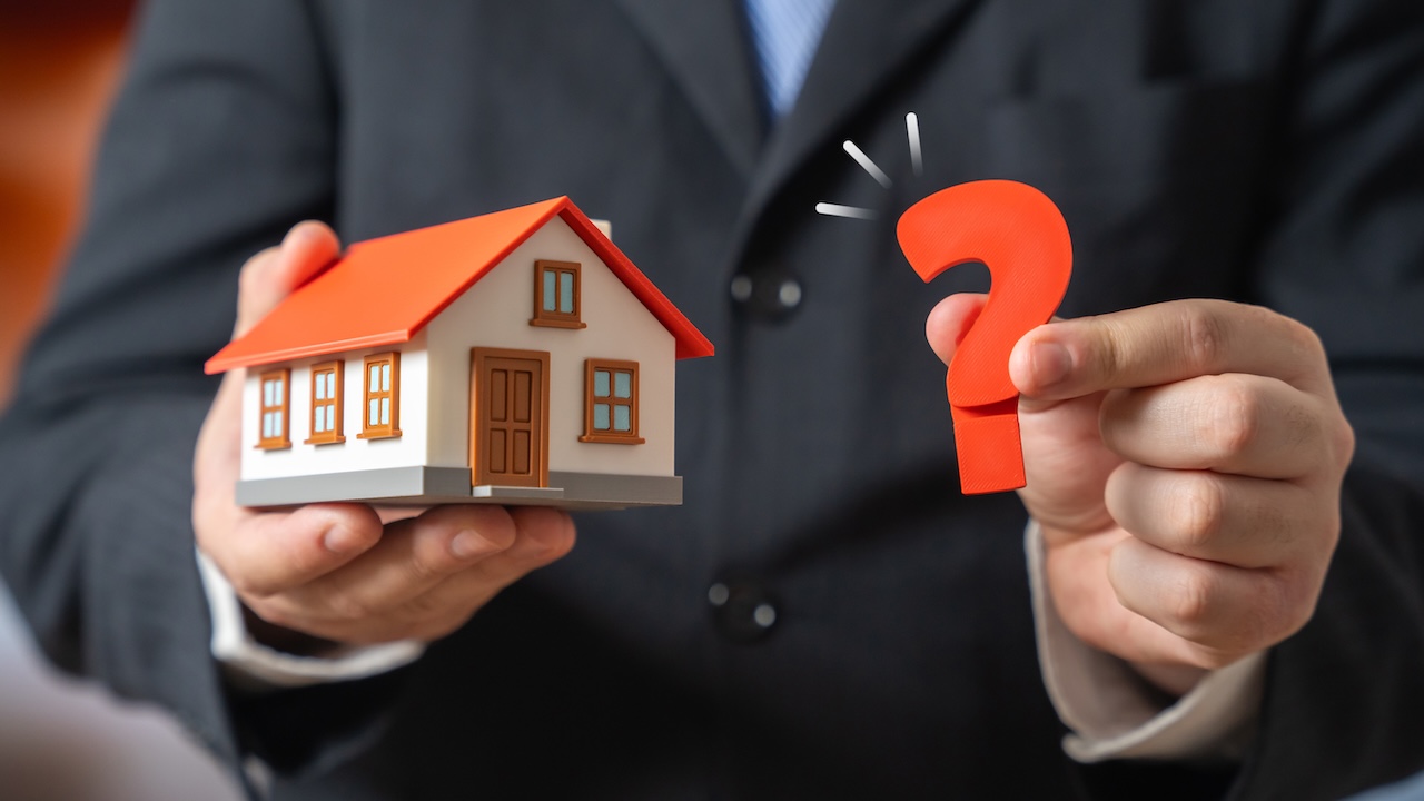 Mann im Anzug hält in den Händen ein Miniaturhaus und ein Fragezeichen