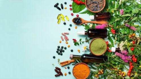 Verschiedene Nahrungsergänzungsmittel wie Kapseln, Tabletten, Pulver und Flüssigpräparate liegen neben Kräutern, Beeren und Heilpflanzen auf hellblauem Hintergrund.