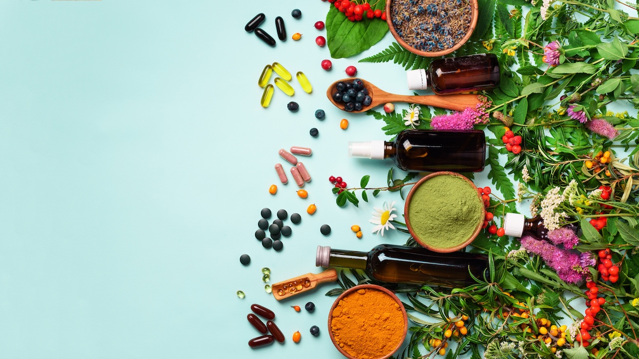 Verschiedene Nahrungsergänzungsmittel wie Kapseln, Tabletten, Pulver und Flüssigpräparate liegen neben Kräutern, Beeren und Heilpflanzen auf hellblauem Hintergrund.