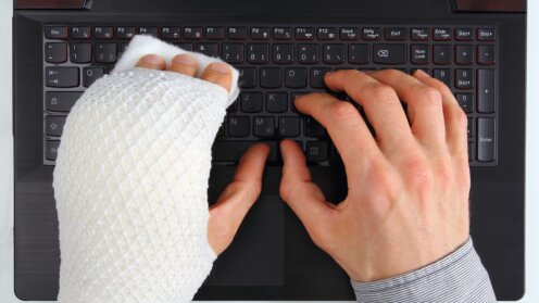 Person mit bandagierter Hand tippt mit der unverletzten Hand auf einer Laptop-Tastatur.
