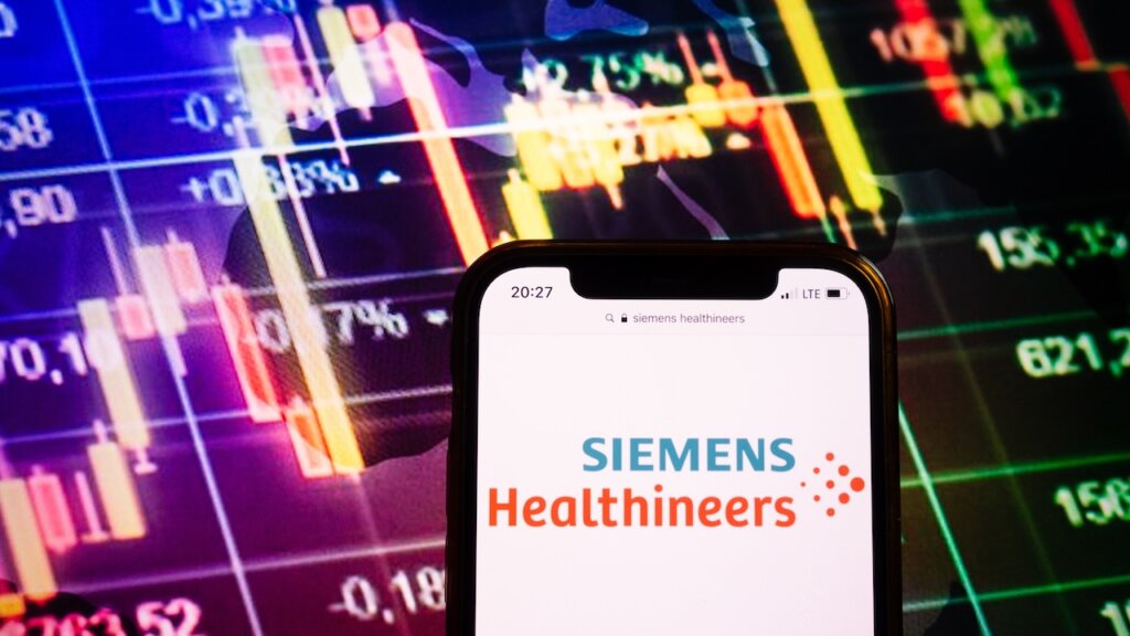 Smartphone mit dem Logo von Siemens Healthineers vor einem Hintergrund mit Börsenkursen und Candlestick-Charts.