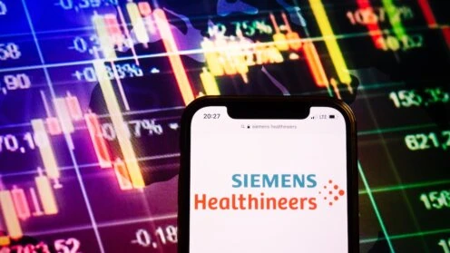 Smartphone mit dem Logo von Siemens Healthineers vor einem Hintergrund mit Börsenkursen und Candlestick-Charts.